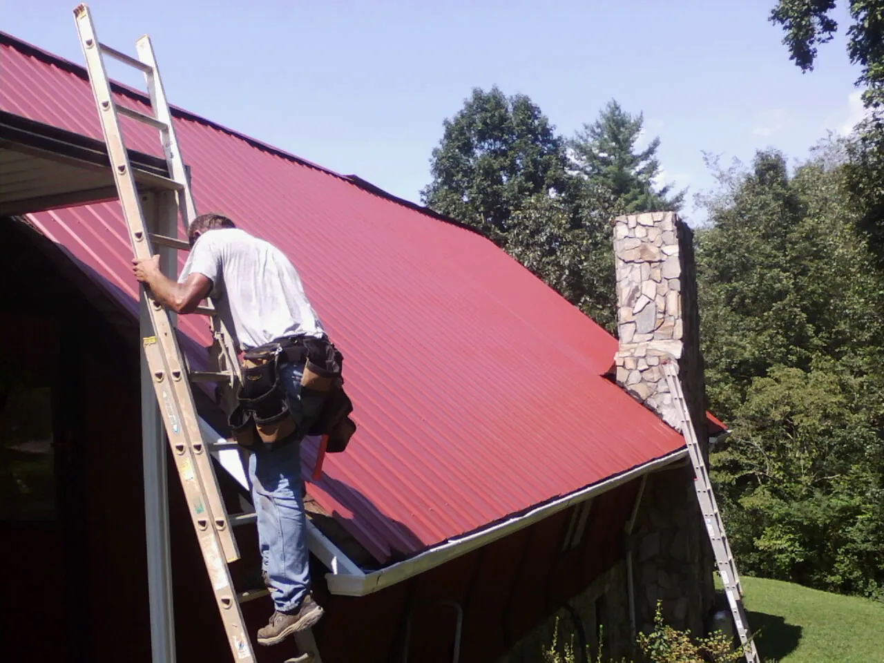 Roofer on a ladder beside a red metal roof and stone chimney — Soffit & Fascia Repair by Roofing Brothers in Worley